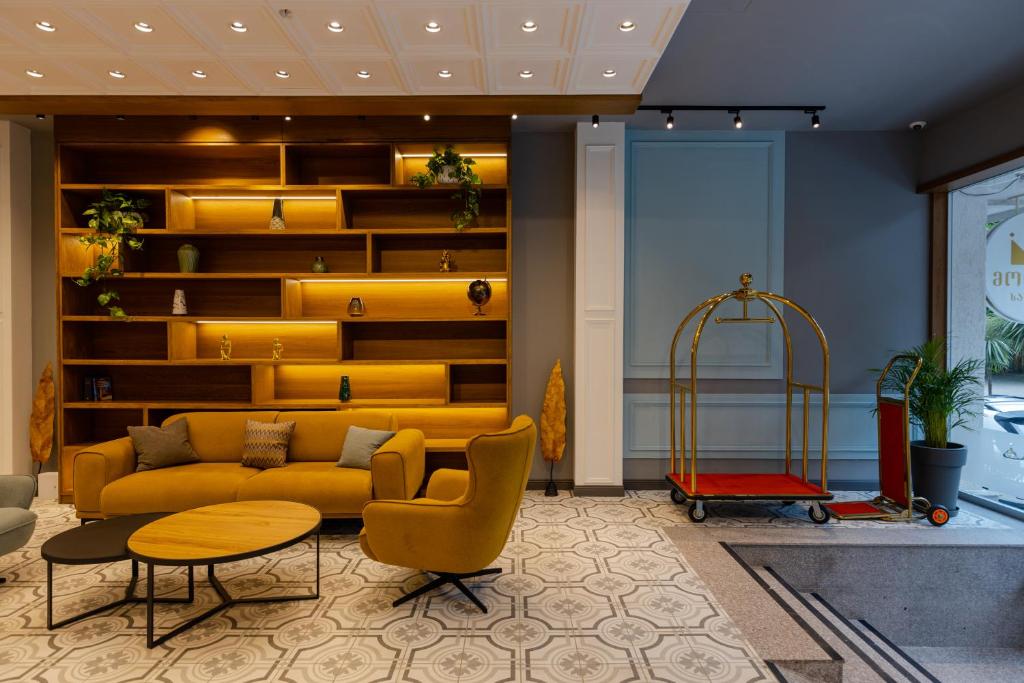 Mūsdienīgs viesnīcas interjers ar dzeltenu dīvānu, melnu galdiņu un stilīgu plauktu sienu, kas izgaismota ar siltām gaismām. Redzams arī krāšņs bagāžas ratiņš un dekoratīvas augu kompozīcijas.