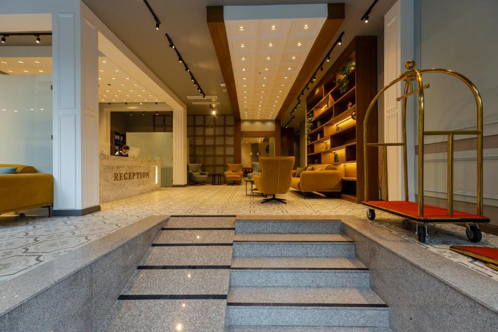 Mūsdienīgs viesnīcas reģistratūras interjers ar stilīgām mēbelēm un ērtiem sēdekļiem, kas rada viesmīlīgu atmosfēru. Skats uz reģistratūras letni un gaidīšanas zonu ar dekoratīvām plauktiem.