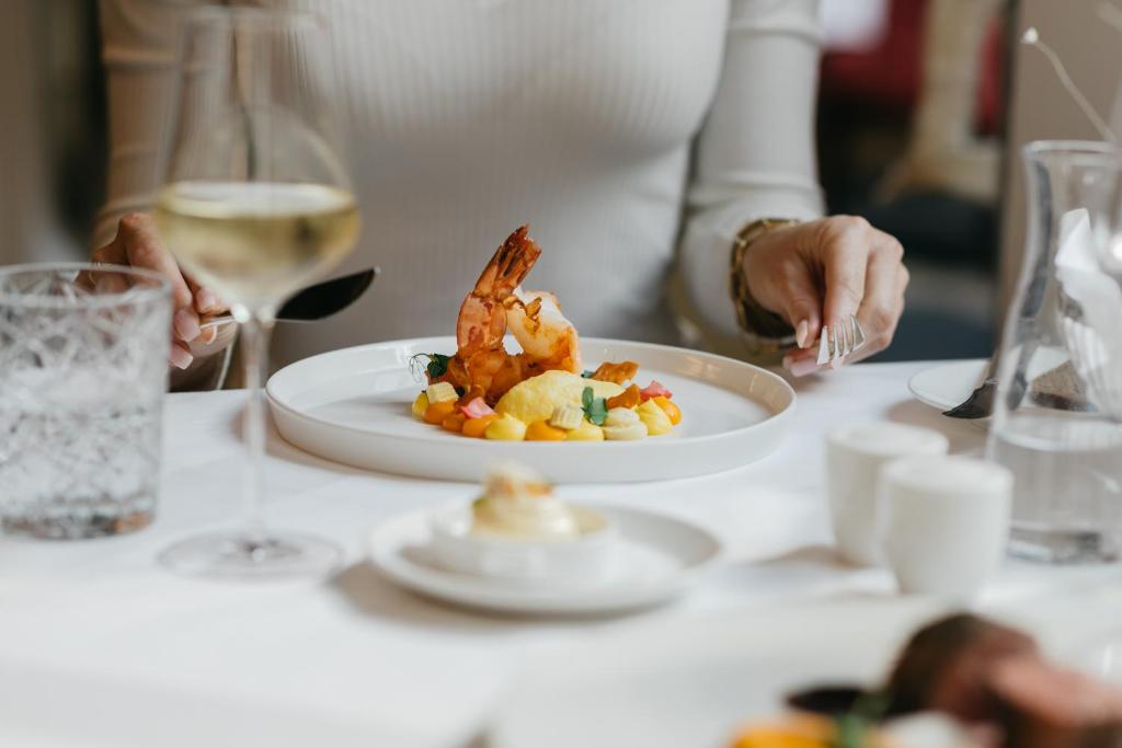 Eleganta restorāna maltīte ar garnelēm un krāsainām dārzeņu garnējumiem, ko pasniedz kopā ar baltvīnu. Galda klājums ir smalks, radot patīkamu atmosfēru.