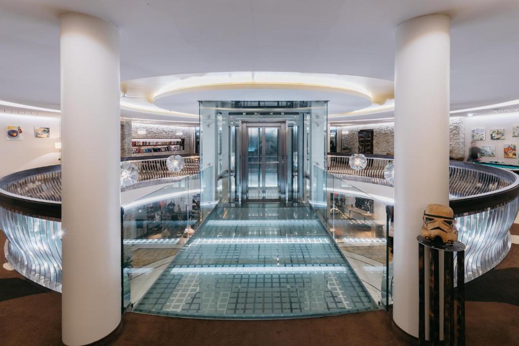 Mūsdienīga un eleganta interjera skats ar stikla liftu un rotājošām lampām, kas rada gaisīgu atmosfēru. Telpa ir aprīkota ar mākslas darbiem un unikālām dekorācijām.