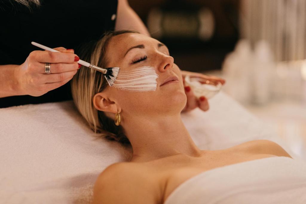 Sieviete saņem sejas procedūru, kurā estēte uzklāj masku ar otu. Fons ir relaksējošs, ar dažādām kosmetikas pudelēm.