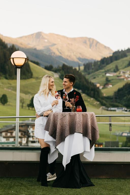 Romantisks brīdis pāris, kas svin dzīves priekus ar dzērieniem uz terases, fona kalni un ainava.