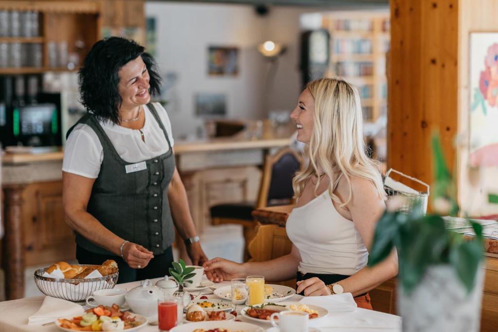 Siltajā restorānā viesmīle pasniedz brokastis apmeklētājai, kurai ir priecīga seja. Galda klājumā redzama dažāda veida ēdienu izvēle, tostarp augļi un dzērieni.