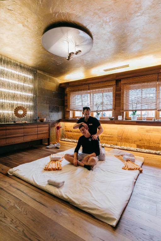 Masāžas sesija relaksējošā vidē ar vīrieti uz paklāja un masieri aiz muguras. Interjers ir elegants ar dabīgiem materiāliem un mierīgu apgaismojumu.