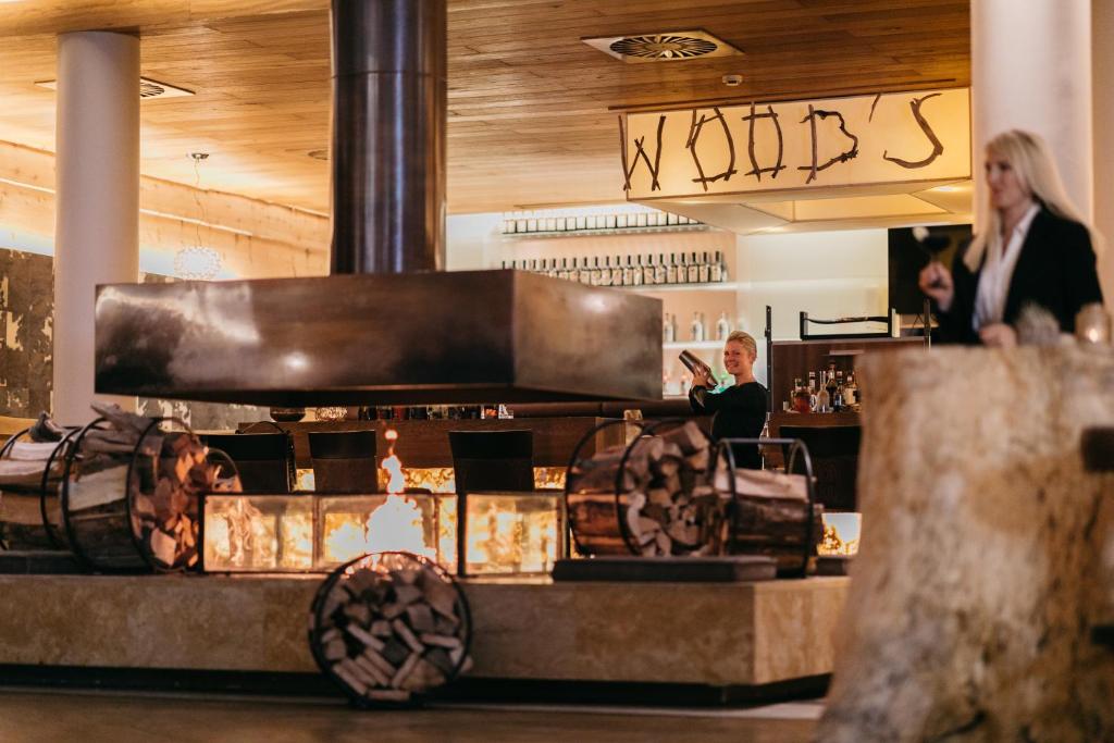 Mūsdienīga bāra iekārtojums ar centrālo kamīnu un koka detaļām, kur apkalpojošs personāls sagatavo dzērienus. Gaismas un siltuma atmosfēra rada patīkamu noskaņu.