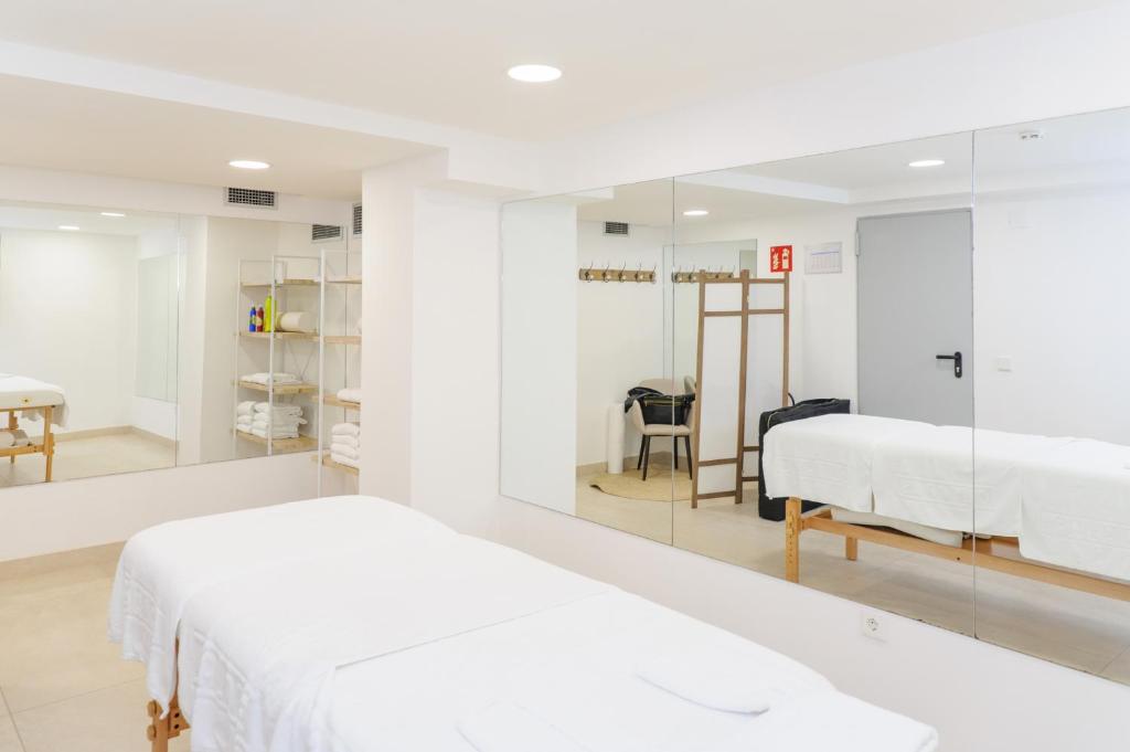 Relaksējošs masāžas telpas interjers ar divām masāžas galdiēm, spoguļiem un plauktiem ar dvieļiem un masāžas piederumiem. Telpa ir gaiša un moderna, radot mierīgu atmosfēru.