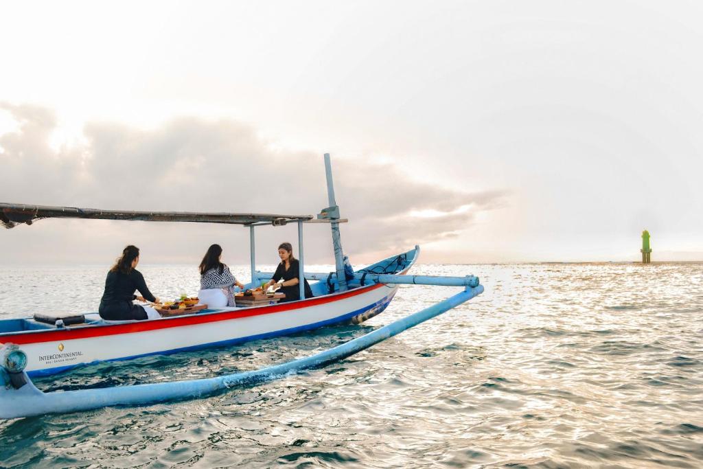 Trīs sievietes gatavo ēdienu uz laivas, kas peld pa mierīgu jūru, ar gaismu, kas atspoguļojas ūdenī. Tālumā redzams zāles bāka.