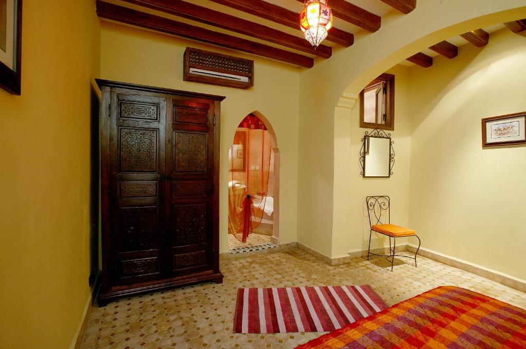 Interjera attēls ar koka skapīti, siltu dzeltenu krāsu un tradicionālu apgaismojumu, radot mājīgu atmosfēru. Gultas veļa ir ar siltām krāsām, un telpā ir redzami dekoratīvi elementi.