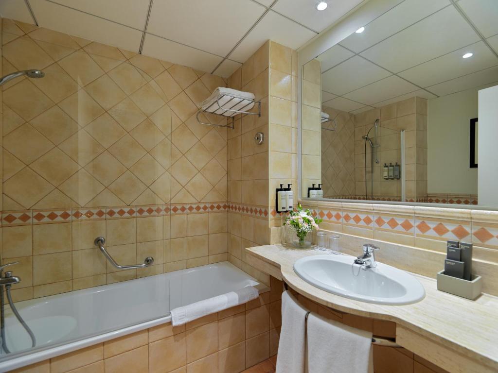 Mūsdienīgs vannas istabas interjers ar vannu, dušu un elegantu izlietni, apgaismojums un flīžu apdare rada patīkamu atmosfēru. Uz galda ir ziedi un higiēnas piederumi.