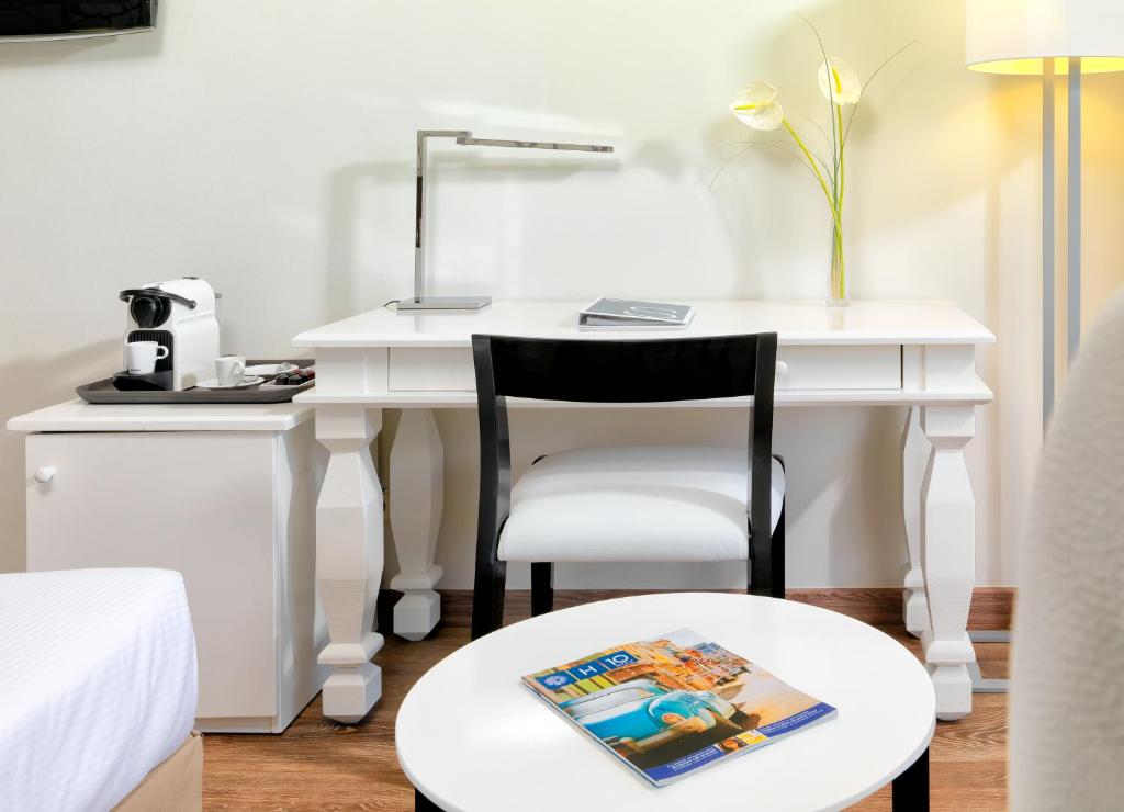 Gaiša viesnīcas istaba ar baltu galdu, melnu krēslu un kafijas automātu. Uz galda atrodas grāmata un lampa, radot mājīgu atmosfēru.