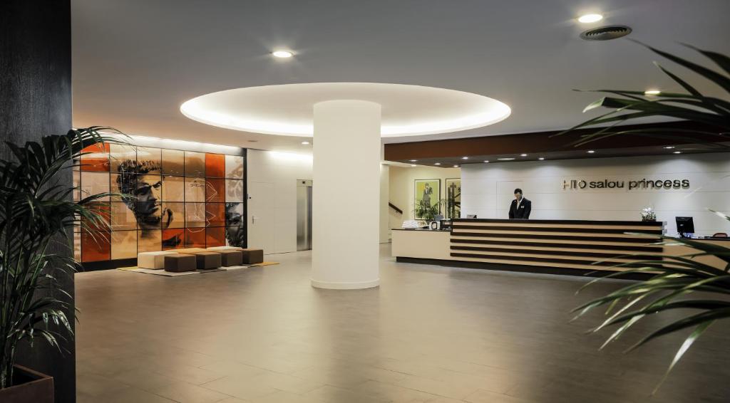 Mūsdienīga viesnīcas recepcija ar elegantu dizainu, kurā redzams mākslas darbs uz sienas un viesmīlis pie reģistratūras. Interjers ir gaišs un atvērts, radot patīkamu atmosfēru.