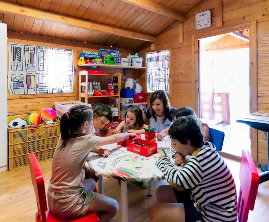 Bērni un pieaugušais radoši strādā pie galda, zīmējot un krāsojot bērnu istabā, kas ir piepildīta ar rotaļlietām un radošiem materiāliem. Gaisotne ir draudzīga un radoša.