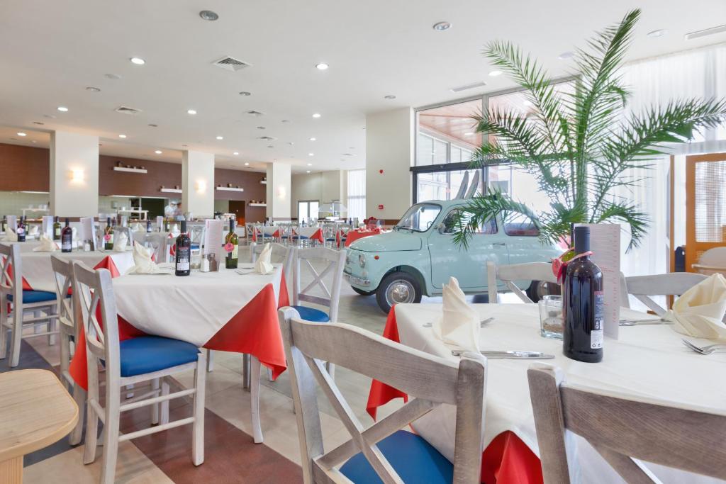 Restorāna iekšskats ar gaišu, modernu dizainu un raksturīgu vintage automobili, kas novietots blakus ēdamgaldiem ar sarkaniem un baltiem galdautiem. Interjerā ir redzami augi un viesmīlīga atmosfēra.