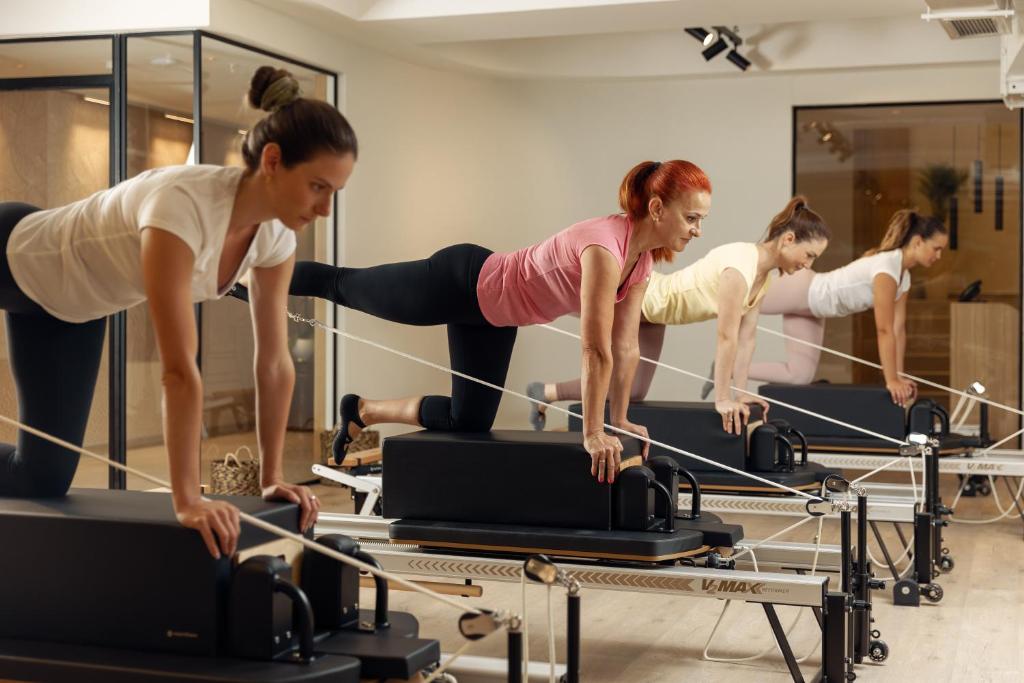 Trīs sievietes nodarbojas ar pilates treniņu uz reformatora, koncentrējoties uz stiepšanās un spēka vingrinājumiem. Interjers ir moderns un gaišs, radot motivējošu atmosfēru.