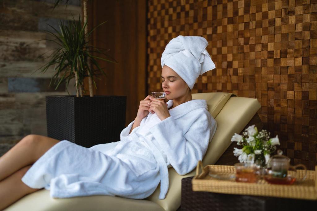 Relaksējoša brīvdiena spa, sieviete baltā halātā un dvielī, baudot dzērienu. Apkārt ir augi un ziedi, kas rada mierīgu atmosfēru.