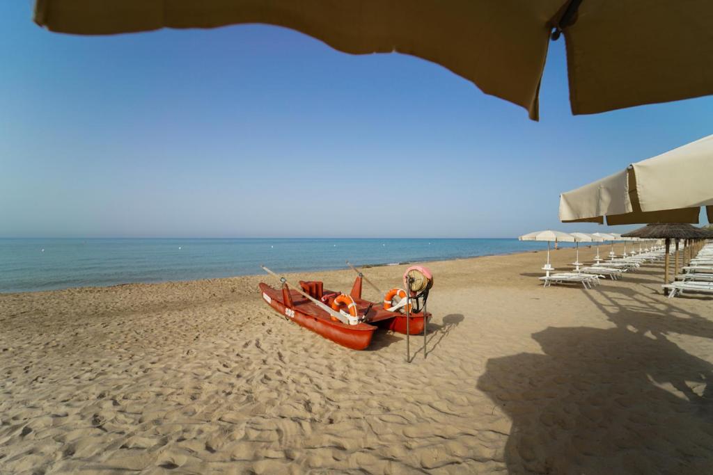 Sarkans laivas uz pludmales smiltīm, blakus saulessargi un atpūtas krēsli, mierīga jūra un skaidrs debesis.