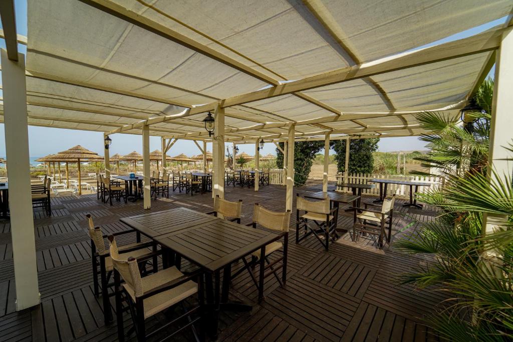 Atvērts restorāna terases skats ar koka galdiem un krēsliem, kas izvietoti zem nojumes. Redzama pludmales atmosfēra un zaļumi fonā.