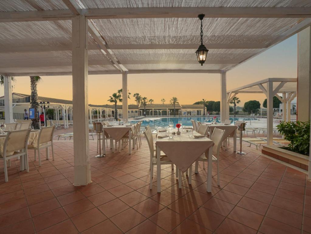 Terases restorāns ar galdiem un krēsliem, kas vērsti uz baseinu, saulrieta gaisma rada relaksējošu atmosfēru. Apzaļumojums un palmas papildina viesnīcas vidi.