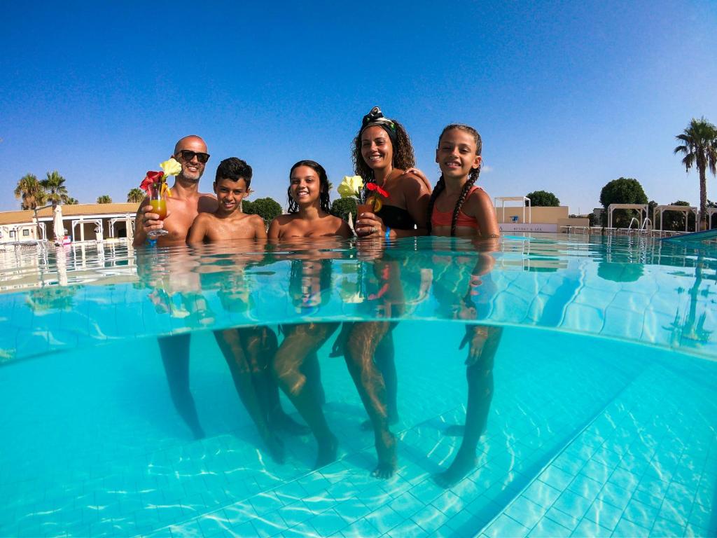 Vasaras brīvdienu mirklis pie baseina, kur pieci cilvēki pozē ar ziediem rokās. Ūdens ir dzidrs un saulains laiks rada priecīgu atmosfēru.