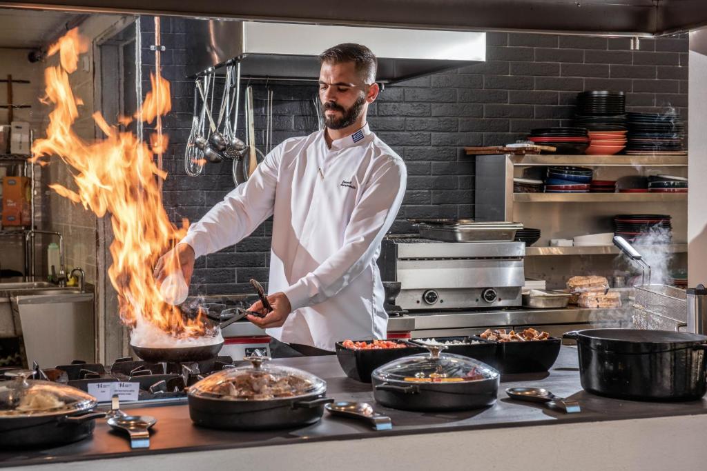 Šefpavārs, kas strādā modernā virtuvē, veicot flambe tehniku ar liesmu, apstrādājot ēdienus. Priekšplānā redzami dažādi ēdieni, kas gatavoti uz galda.