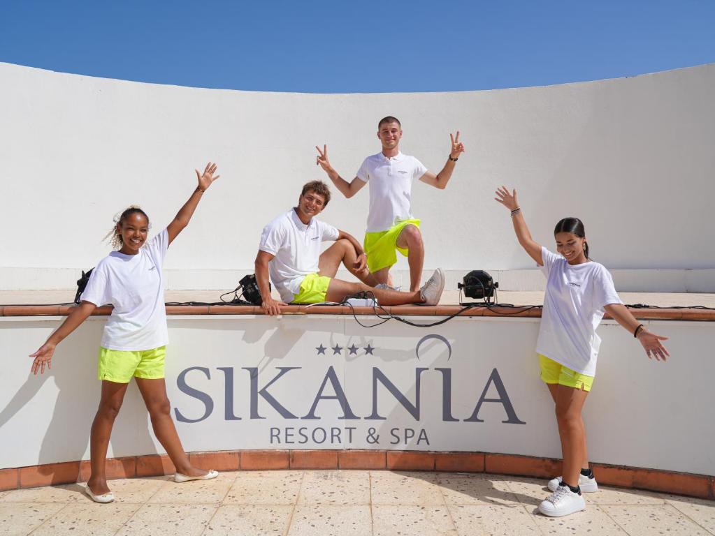 Četri cilvēki jautrā pozā pie Sikania Resort & Spa logo, ģērbti baltās t-kreklos un spilgti dzeltenās šortos, ar sauli un skaidru debesi fona.