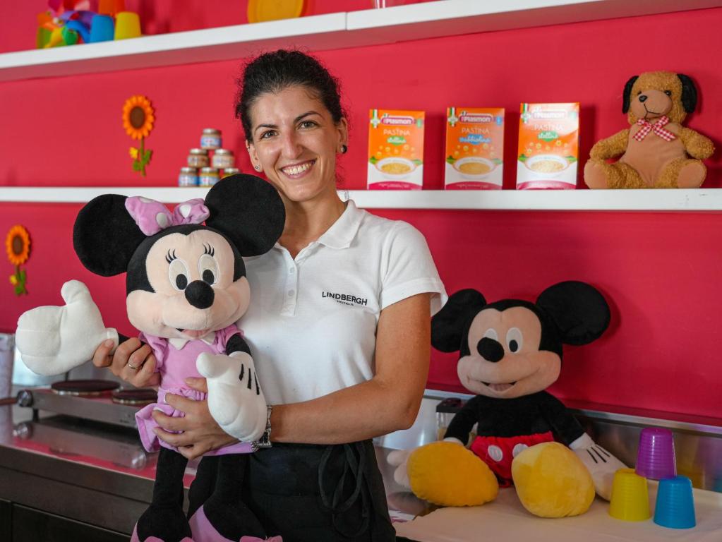 Smaidoša sieviete pozē ar lielu Minnie Mouse lelli, kamēr fona ir krāsains un bērnu rotaļlietas. Stends ir dekorēts ar saulītēm un bērnu ēdieniem.