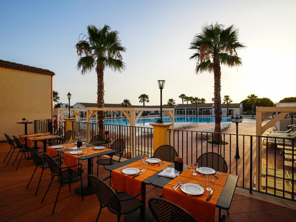 Restorāna terase ar skatu uz baseinu, kur galdi ar oranžiem galdautiem ir sagatavoti viesu uzņemšanai. Apkārt aug palmām un noriet saule.