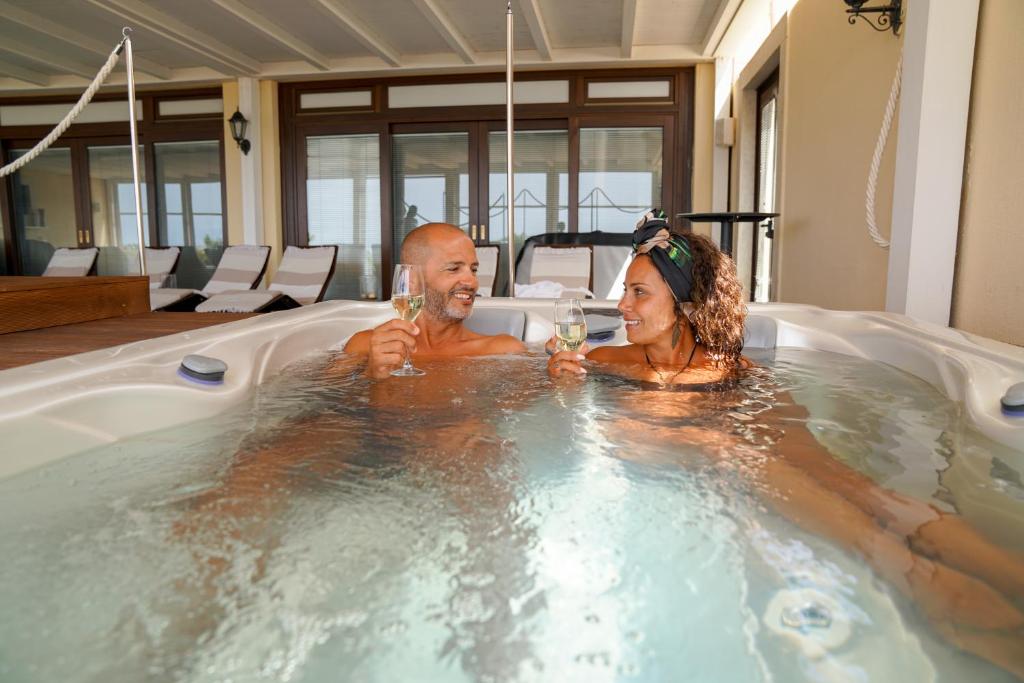 Pāris relaksējas džakuzi, turot rokās šampanieša glāzes un baudot romantisku brīdi. Fons ar lieliem logiem un sauļošanās krēsliem veido patīkamu atmosfēru.