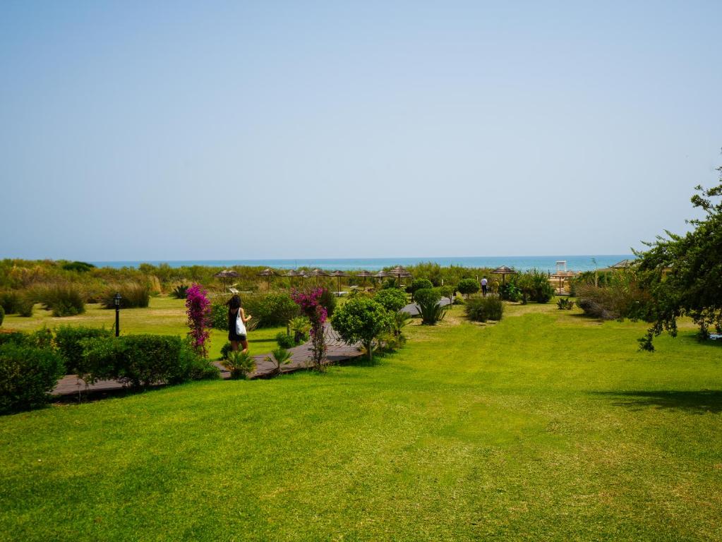 Plaša pludmales ainava ar zāli un krūmiem, kas ved uz jūru, kur redzami pludmales saulessargi.