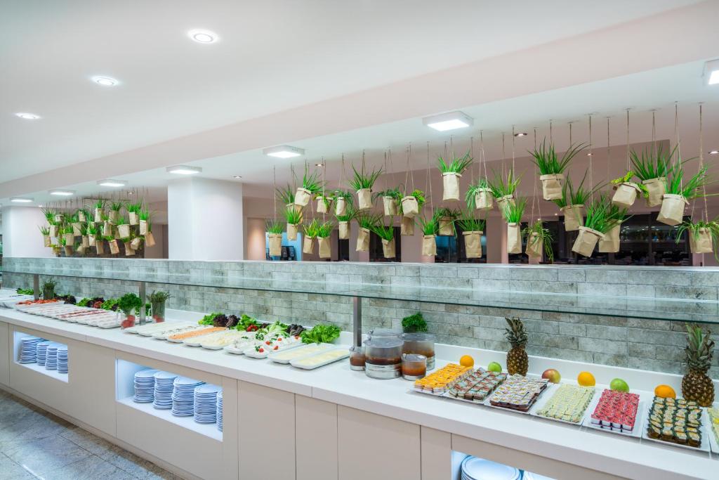 Gaisma un moderns restorāns ar bufeti, kurā redzami dažādi dārzeņi un saldēti ēdieni, kā arī piekārti augi, kas piešķir patīkamu atmosfēru.