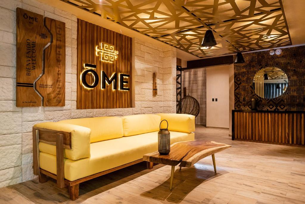 Mūsdienīgs viesnīcas interjers ar dzeltenu dīvānu, koka mēbelēm un stilīgu apgaismojumu. Sienā iekļauts viesmīlības logo un dekoratīvi elementi, kas rada siltu atmosfēru.