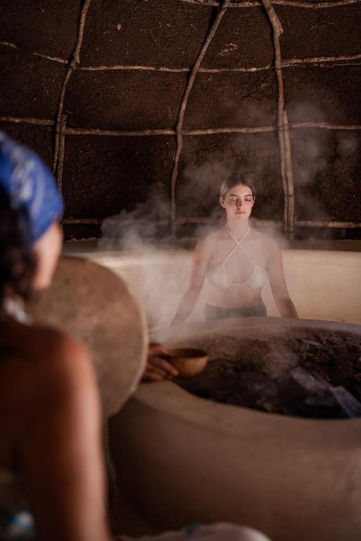 Sieviete meditē tvaika telpā, apkārt valda relaksējoša atmosfēra, kur dūmi paceļas no silta akmens krāsns. Otra sieviete, kas sēž tuvāk, tur rokā mazu trauku.
