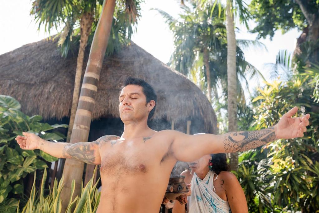 Vīrietis ar tetovējumiem stāv ar izstieptām rokām, baudot dabu un mieru, kamēr fona dārzā ir tropu augi un tradicionāls jumts. Gaisā redzama smaržīga dūmu plūsma.