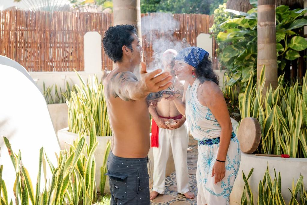 Divi cilvēki piedalās garīgā ceremonijā, kas ietver dūmu izplatīšanu, kamēr trešais cilvēks vēro no fona. Apkārt ir zaļi augi un tropiska vide.