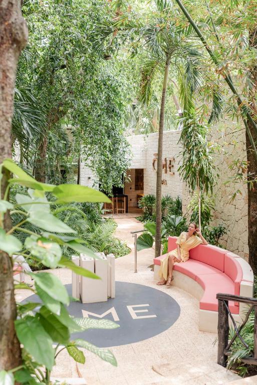 Tropu dārzs ar rozā dīvānu un dabas elementiem, kas rada mierīgu atmosfēru. Fonā redzama ēka ar nosaukumu 