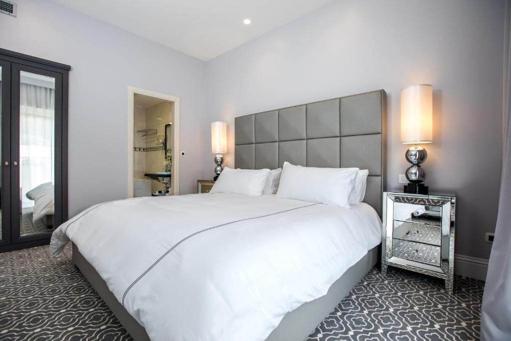 Mūsdienīga guļamistaba ar plašu gultu, elegantu interjeru un spoguļu naktsgaldiņu. Telpa ir gaiša un komfortabla, ar stilīgu paklāju un modernām gaismas ķermeņiem.