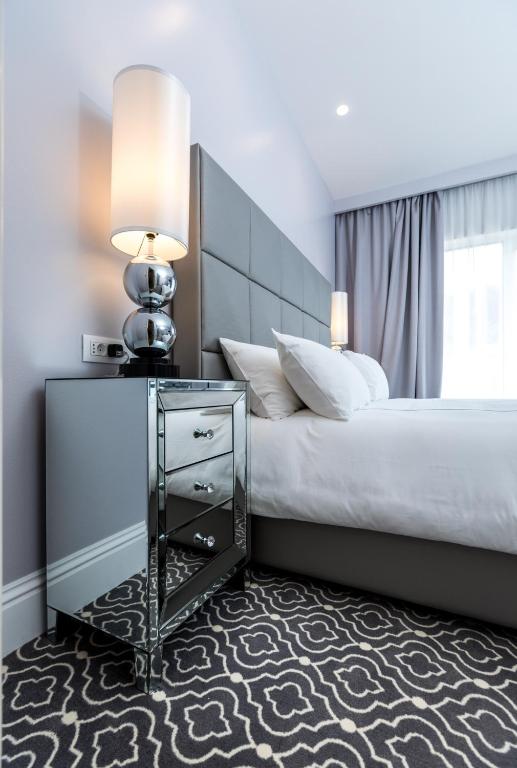 Moderns guļamistabas interjers ar elegantu naktsgaldiņu, stilīgu lampu un mīkstu paklāju. Gaismas toņi un mūsdienīgs dizains rada relaksējošu atmosfēru.