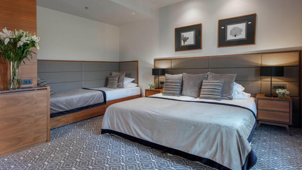 Mūsdienīgs guļamistabas interjers ar diviem gultām, elegantiem gultas piederumiem un dekoratīvām gleznām uz sienas. Telpā ir arī ziedu kompozīcija un stilīgi naktsgaldiņi ar lampām.