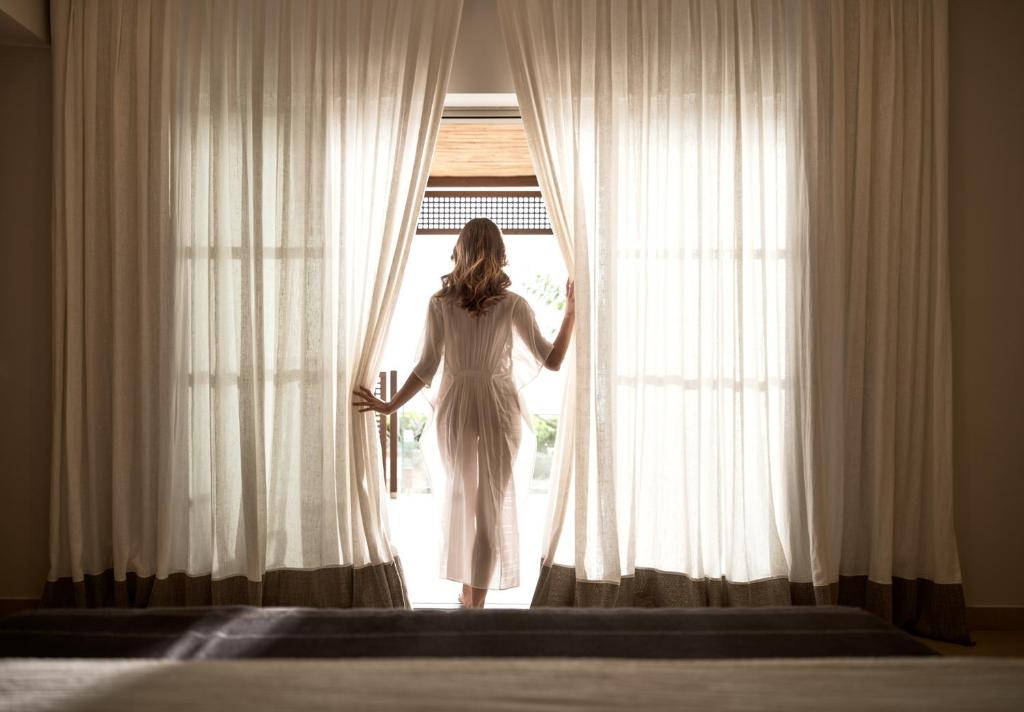 Sieviete baltā halātā stāv pie atvērtām durvīm, iznākot no istabas un raugoties caur plānām aizkariem. Gaisma ieplūst telpā, radot mierīgu un relaksējošu atmosfēru.