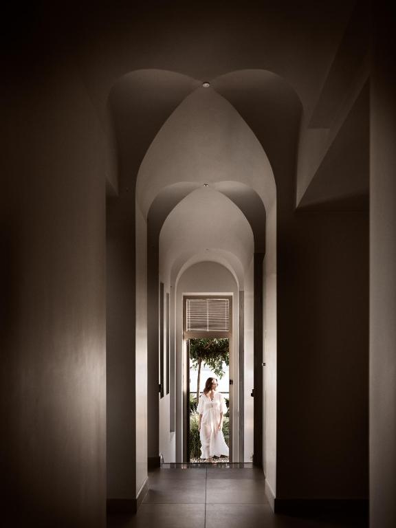 Eleganta un mūsdienīga koridora aina ar arku formas griestiem, kurā redzama sieviete baltā apģērbā, stāvot pie loga, caur kuru ieplūst dabiska gaisma.