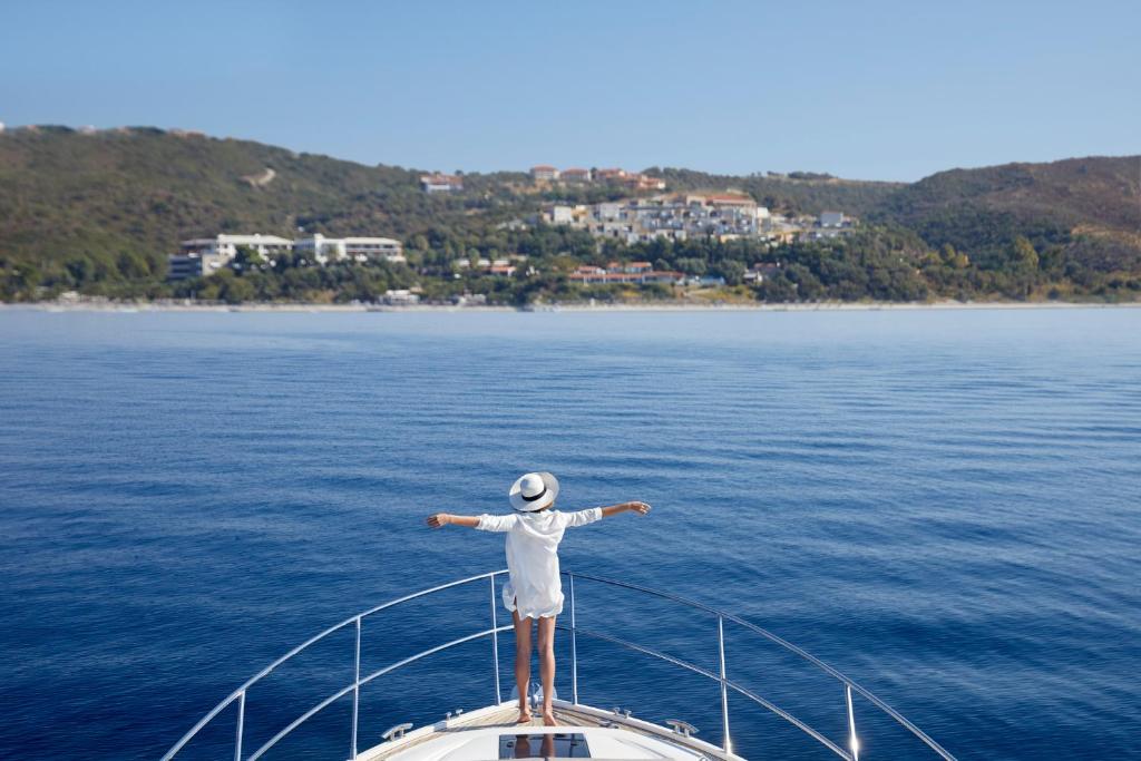 Sieviete stāv uz jahtas priekša, izstiepjot rokas un baudot mierīgu jūras ainavu ar pakalniem un mājām fona.