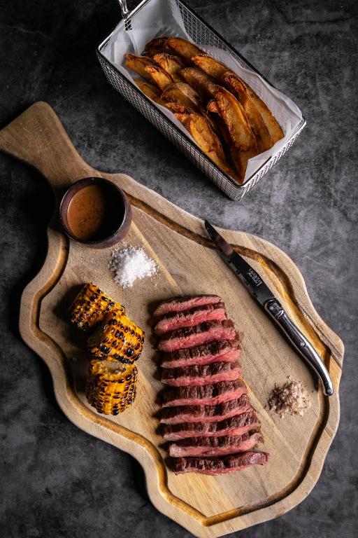 Grilēta gaļa, grauzdēta kukurūza un cepti kartupeļi, pasniegti uz koka griešanas dēļa ar mērci un sāli.