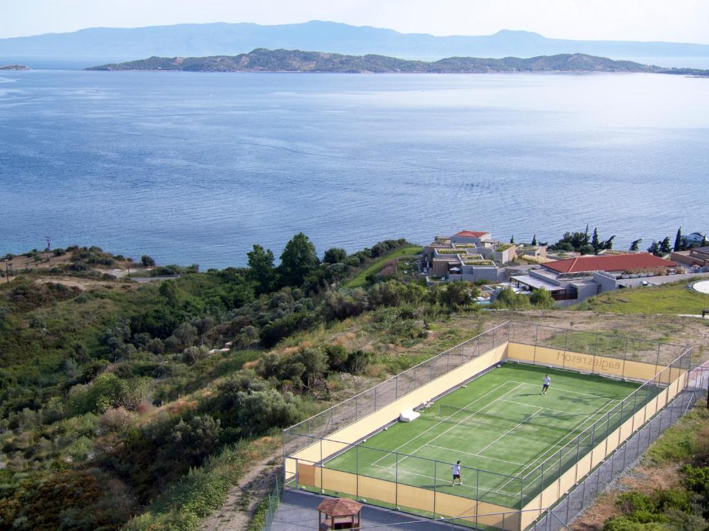 Skats uz tenisa laukumu pie jūras, ar kalniem fona. Laukums ir norobežots, un spēlētāji ir aktīvi spēlē. Apkārt esošā daba un ainava veido mierīgu atmosfēru.