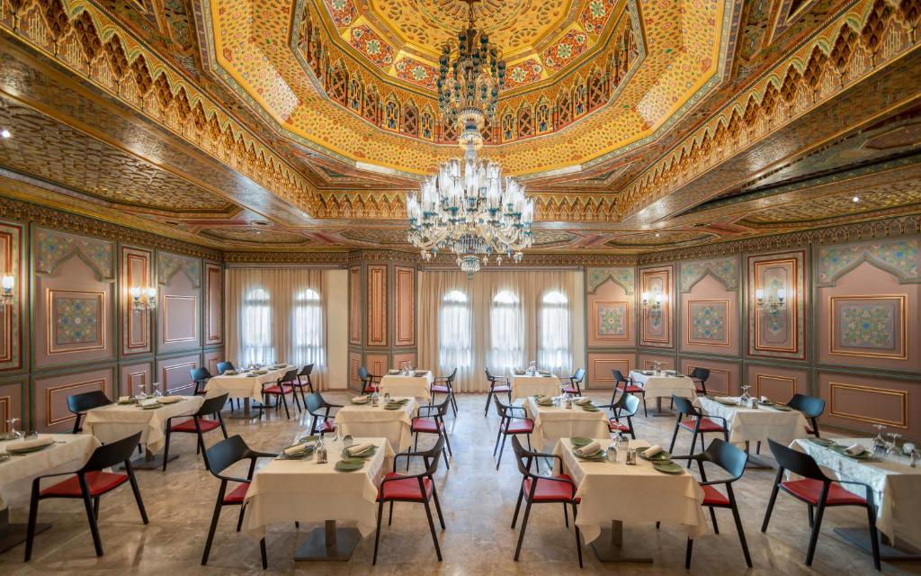 Elegants restorāns ar izsmalcinātu interjeru, rotātu ar krāsainiem griestiem un kristāla lustru. Galdi ir sagatavoti ar galda piederumiem un salvetēm, radot patīkamu atmosfēru.