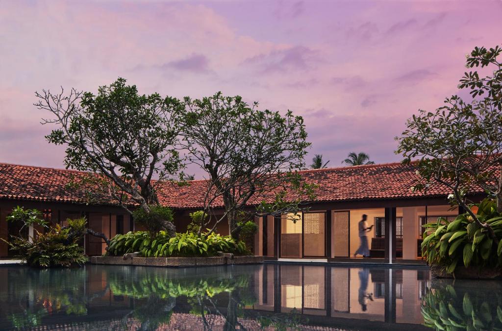 Mūsdienīga viesnīca ar skaistu ainavu, kurā redzami zaļi augi, ūdens baseins un sarkano jumtu ēkas, kas atspoguļojas saulrieta gaismā.