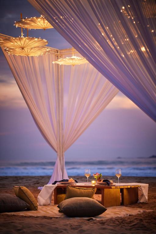 Romantiska pludmales vakariņas zem gaisa laternām, ar gaismiņām un maziem galdiem, kas izkārtoti smiltīs.