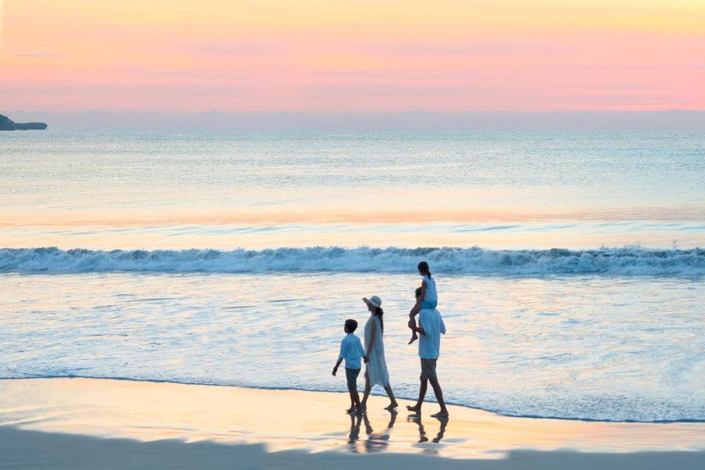 Ģimene pastaigā pie pludmales saulrieta laikā, ar maigu viļņu šalkoņu fonā. Skats uz mierīgu jūru un krāšņu debesu krāsu spēli.