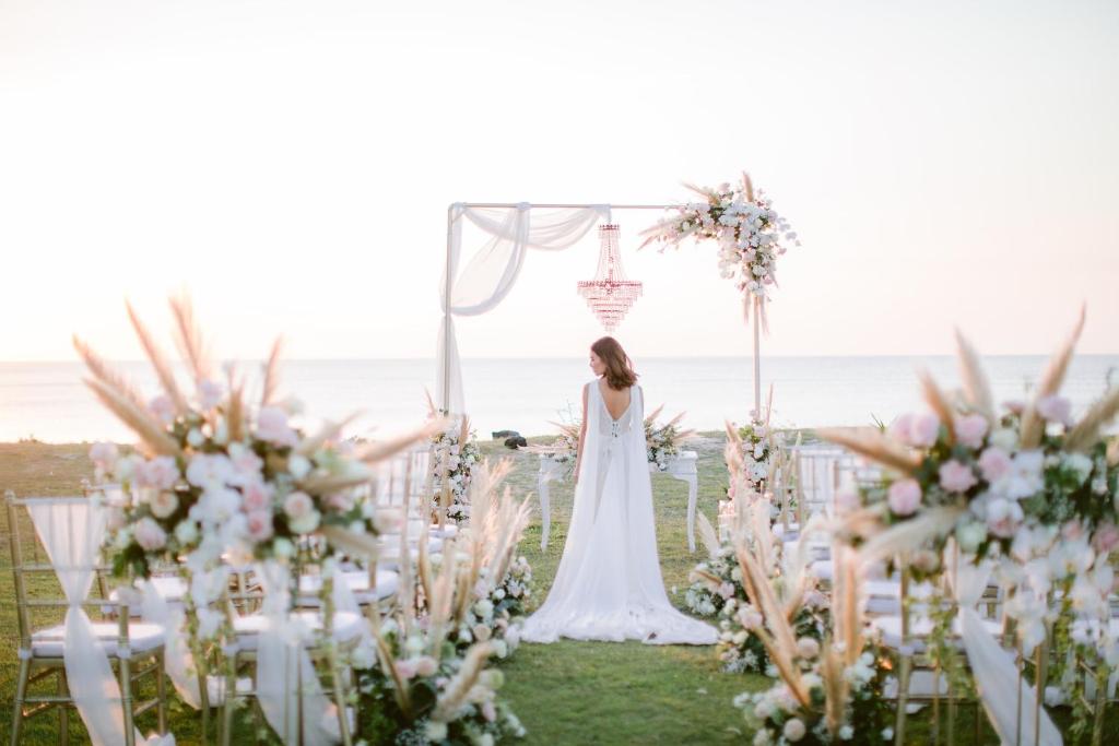 Romantiska pludmales kāzu ceremonija ar ziedu kompozīcijām un dekoratīvām gaismām, kas veido burvīgu atmosfēru saulrieta laikā.