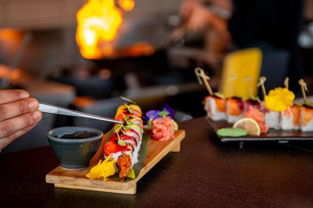 Svaiga sushi pasniegšana ar dekoratīvām sastāvdaļām un garšvielām, kamēr fons ir piepildīts ar kulinārijas aktivitātēm.
