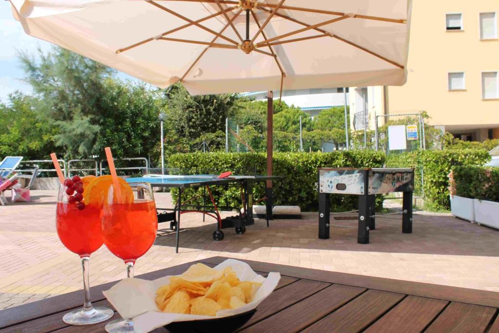 Divas dzirkstošas ​​dziras ar apelsīnu šķēlītēm un ogām, blakus ir čipsi, bet fona ir peldbaseina zona ar galda spēlēm un saulessargu.